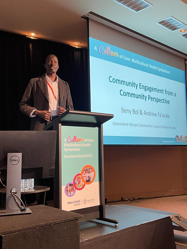 Beny Aterdit Bol speaking from a lectern at a multicultural health symposium 