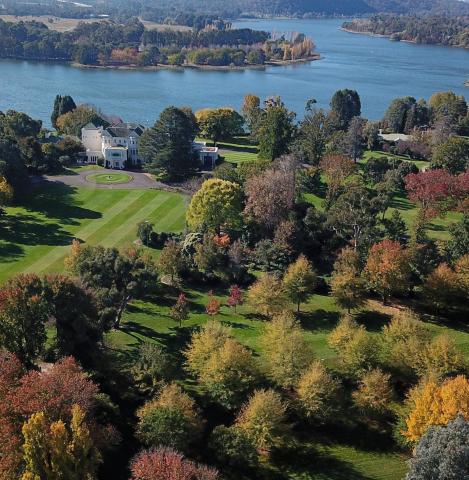 An aerial shot o showing trees and a large white house with a lake behind it