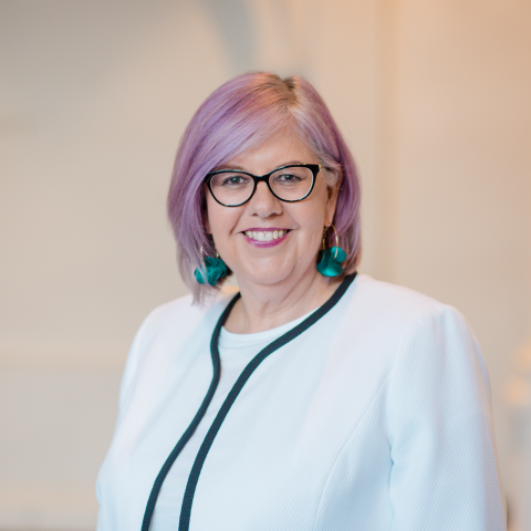 Pia Turcinov AM smiling at camera, wearing white jacket, with purple tinged hair