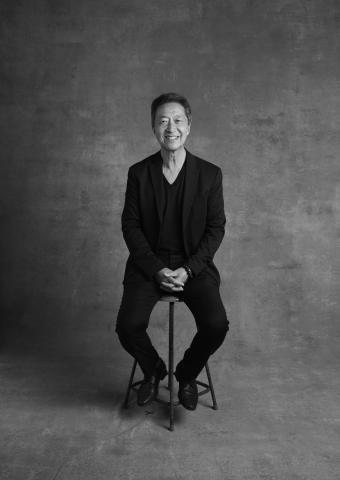 black and white image of Simon Chan sitting on a stool smiling